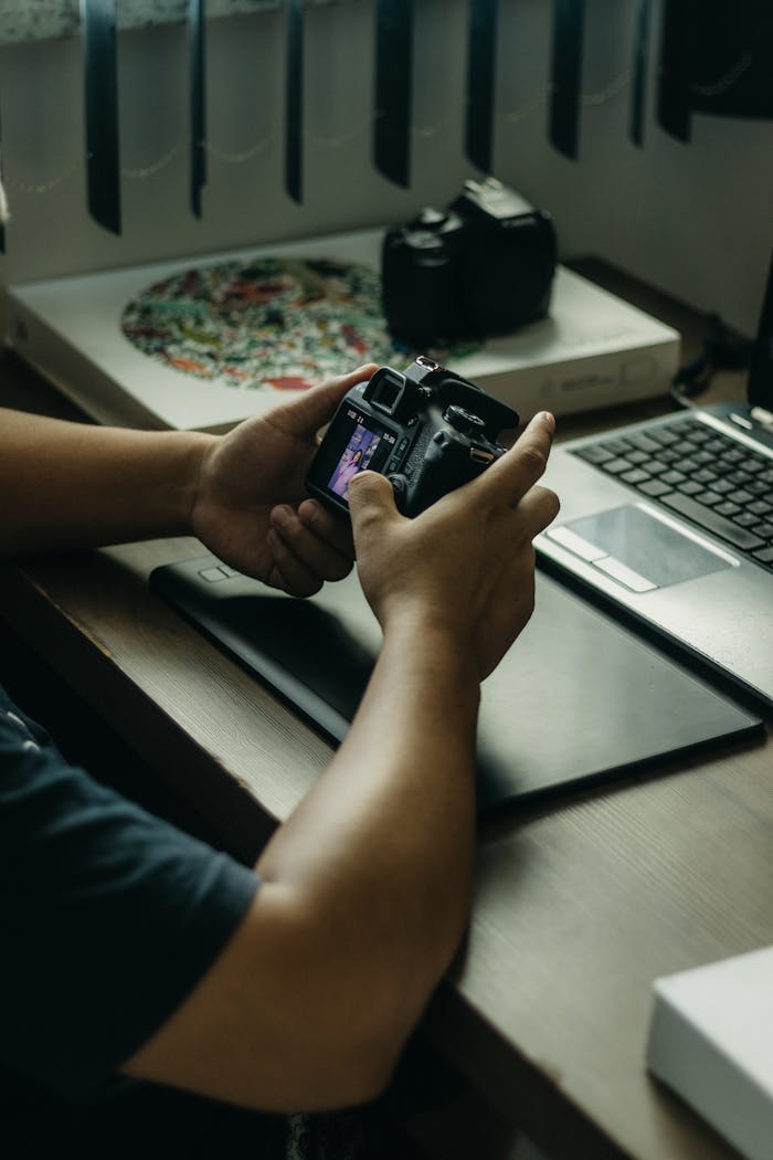 Man holding a camera while working on a laptop, setting up a photography session indoors.